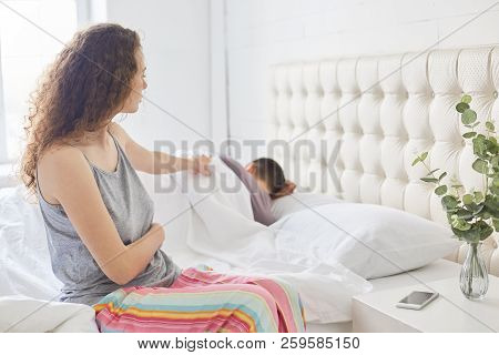 Curly Beautiful Woman In Pyjamas Sits On Side Of Bed, Dressed In Casual Pyjamas, Tries To Awake Her 