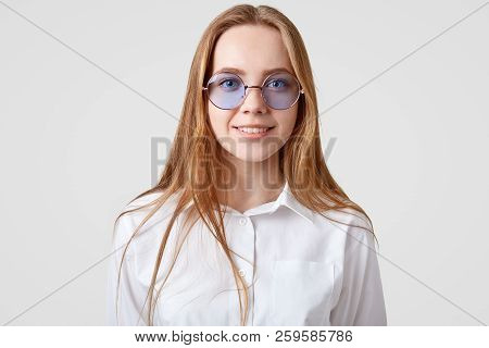 Horizontal Portrait Of Cheerful Female Student Wears White Shirt, Trendy Round Shades, Looks Directl