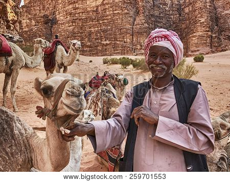 Wadi Rum, Jordan - June 14, 2016: A Smiling Bedouin Camel Herder Holds One Of His Camels In The Cany