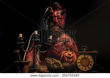 Halloween Devil Man With Pumpkin, Clock, Candlestick On Table. Demon With Horns On Head Sit On Black
