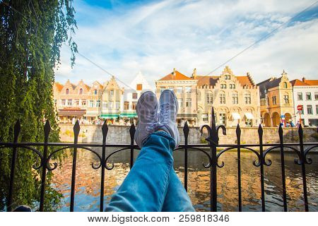 Evening In Brugge. Tourist Legs In White Sneakers On Brugge Cityscape Background. Destination Scene.