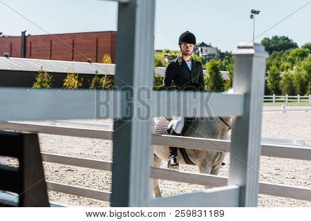 View Through Fence On Handsome Male Equestrian Riding Horse At Horse Club And Looking At Camera