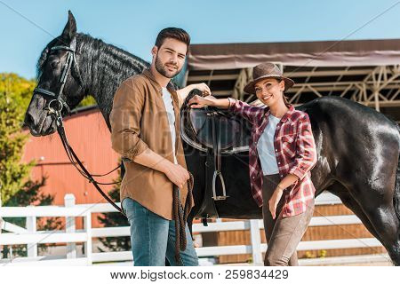 Low Angle View Of Cowboy And Cowgirl Standing Near Horse At Ranch And Looking At Camera