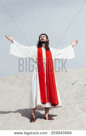 Jesus In Robe, Red Sash And Crown Of Thorns Standing With Raised Hands And Talking With God In Deser