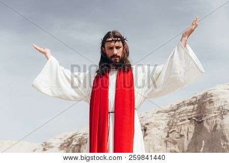 Low Angle View Of Jesus In Robe, Red Sash And Crown Of Thorns Standing With Raised Hands In Desert