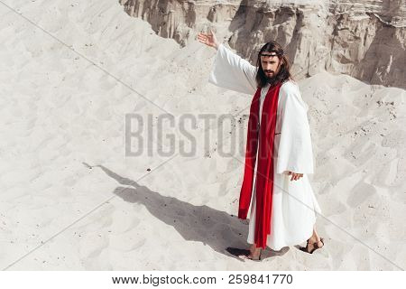 High Angle View Of Jesus In Robe, Red Sash And Crown Of Thorns Showing Way In Desert