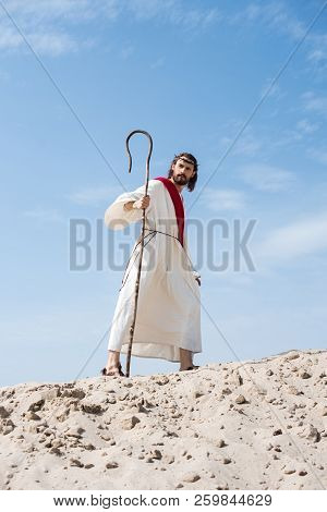 Jesus In Robe, Red Sash And Crown Of Thorns Walking On Sandy Hill With Staff In Desert