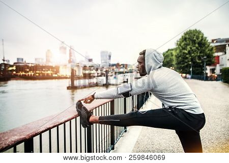 Black Man Runner With Smartphone In An Armband On The Bridge In A City, Stretching.