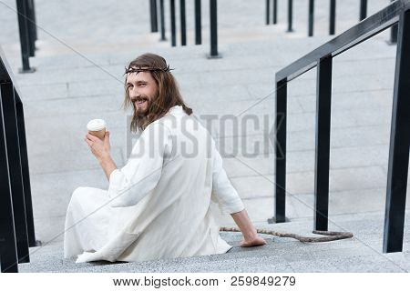 Back View Of Smiling Jesus In Robe And Crown Of Thorns Sitting On Stairs And Holding Disposable Coff