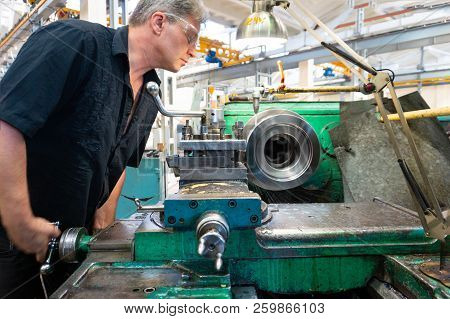 A Worker, A Man In A Black Shirt And Goggles, Controls A Mechanical Machine. Turning Work In Product