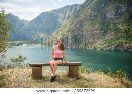 Beautiful Young Woman Looking At Camera While Sitting On Wooden Bench Near Aurlandsfjord And Majesti