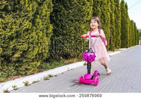 Horizontal Shot Of Happy Pretty Little Girl Wearing Pink Dress And Backapack On The Scooter Against 