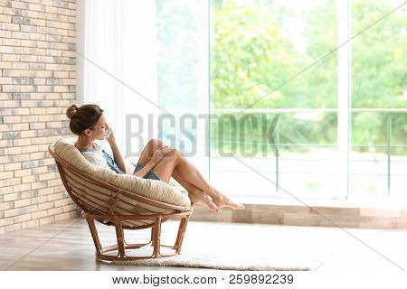 Young Woman With Mobile Phone Sitting In Papasan Chair Near Window At Home