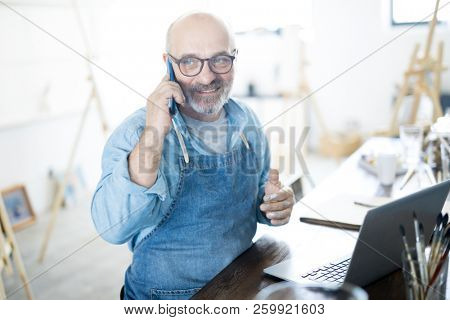 Mature bearded man talking by smartphone while sitting by table in studio and networking