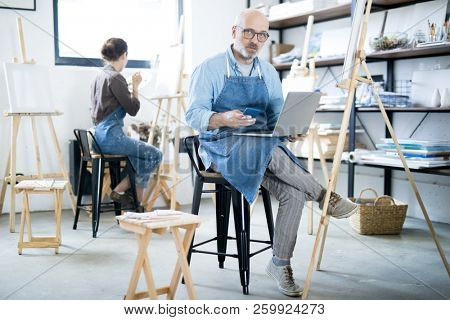 Serious aged man in workwear using modern gadgets while working in studio of arts
