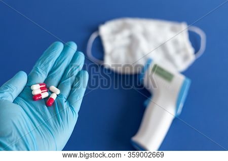 A Hand In A Medical Glove Holds Pills In The Hand, In The Background Lies A Thermometer And A Medica