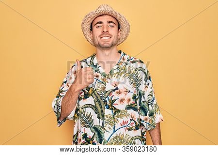 Young handsome man with blue eyes on vacation wearing summer florar shirt and hat doing happy thumbs up gesture with hand. Approving expression looking at the camera showing success.