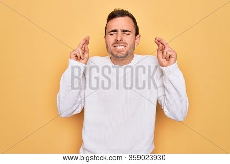 Young handsome man with blue eyes wearing casual sweater standing over yellow background gesturing finger crossed smiling with hope and eyes closed. Luck and superstitious concept.