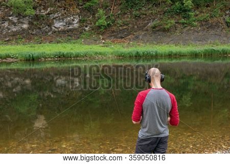 Rear View Of Bald Young Man Standing On River Bank And Listening To Music In Headphones. Far From Bi
