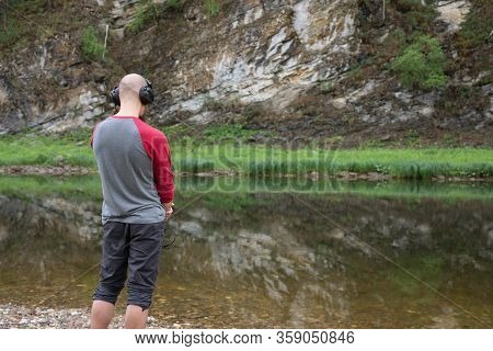 Rear View Of Bald Young Man Standing On River Bank And Listening To Music In Headphones. Far From Bi
