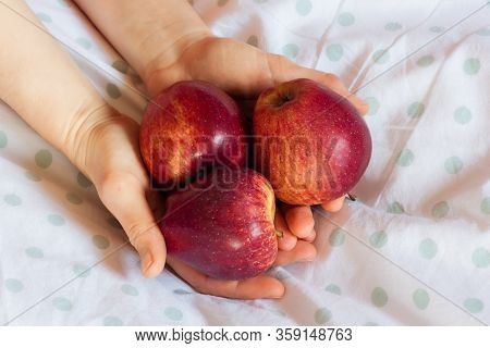 Three Red Apples In The Hands Of A Girl On White Duvet Cover Background On The Bed. Vitamins To Figh