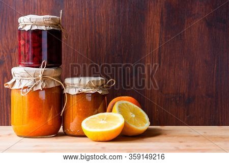Jars With Various Jams And Lemon Cut And Orange On Dark Wooden Background. Strowberry And Apricot Ja