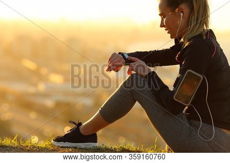 Runner Wearing Earphones Checking Smartwatch Sitting On The Ground At Sunset