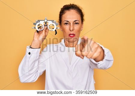 Middle age senior oculist woman holding optometrical glasses for vision correction pointing with finger to the camera and to you, hand sign, positive and confident gesture from the front