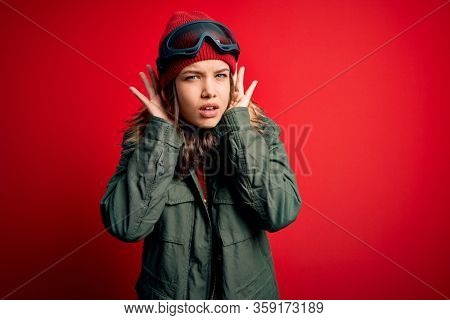 Young blonde girl wearing ski glasses and winter coat for ski weather over red background Trying to hear both hands on ear gesture, curious for gossip. Hearing problem, deaf