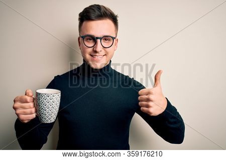 Young handsome caucasian man wearing glasses drinking a cup of hot coffee happy with big smile doing ok sign, thumb up with fingers, excellent sign