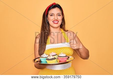 Young hispanic pin up woman wearing fashion sexy 50s style holding sweet cupcakes happy with big smile doing ok sign, thumb up with fingers, excellent sign