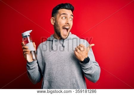 Young handsome sportsman drinking bottle of water to refeshment over red background pointing and showing with thumb up to the side with happy face smiling
