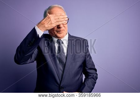 Grey haired senior business man wearing glasses and elegant suit and tie over purple background covering eyes with hand, looking serious and sad. Sightless, hiding and rejection concept