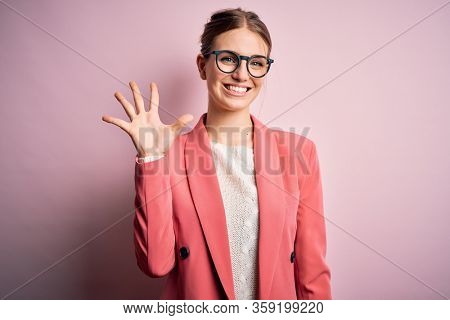 Young beautiful redhead woman wearing jacket and glasses over isolated pink background showing and pointing up with fingers number five while smiling confident and happy.