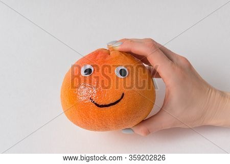 Hand Holding Big Orange Smiley On White Background. Grapefruit With Googly Eyes And Smile.