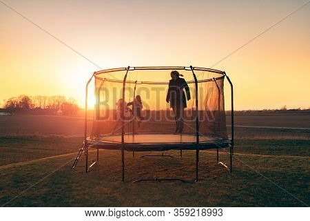 Trampoline On A Lawn In The Sunset With Two Kids And A Young Woman Juping And Playing