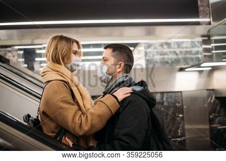 Side View Of Hugging Couple In Medical Masks On Escalator In Subway. Coronavirus Pandemic.