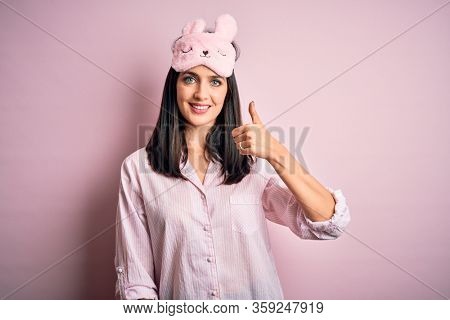 Young brunette woman with blue eyes wearing pink pajama and eye mask over pink background happy with big smile doing ok sign, thumb up with fingers, excellent sign