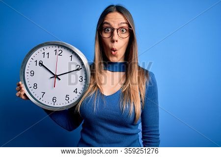 Young blonde woman with blue eyes holding big minute clock over isolated background scared in shock with a surprise face, afraid and excited with fear expression