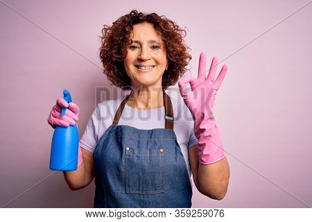 Middle age curly hair woman cleaning doing housework wearing apron and gloves using spayer doing ok sign with fingers, excellent symbol