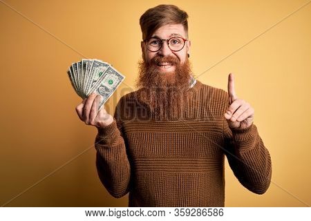 Redhead Irish man with beard holding a bunch of 1 dolla banknotes over yellow background surprised with an idea or question pointing finger with happy face, number one