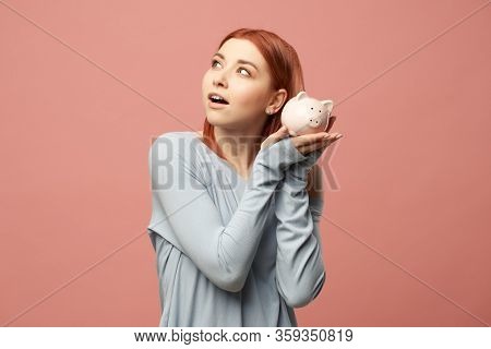 Young ginger woman with piggy bank in hands standing in studio