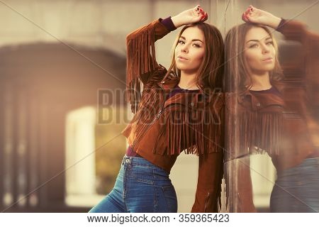 Happy young fashion woman leaning on the wall Stylish female model in brown leather fringe suede jacket and dark blue jeans