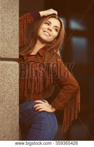 Happy young fashion woman leaning on the wall Stylish female model in brown leather fringe suede jacket and dark blue jeans