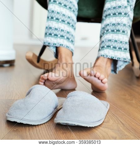 closeup of a man in pajamas, sitting on a modern rocker, about to put on a pair of gray slippers placed on the laminate floor in the foreground