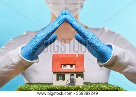 Woman In Medical Gloves And Mask Holds Hands Over A House. Concept Of Home Stay, Quarantine, Securit