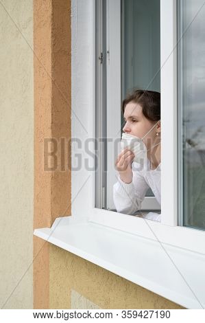 Caucasian Young Woman In Protective Mask Looks Out Of An Open Window. Stay Home Concept. Quarantine 