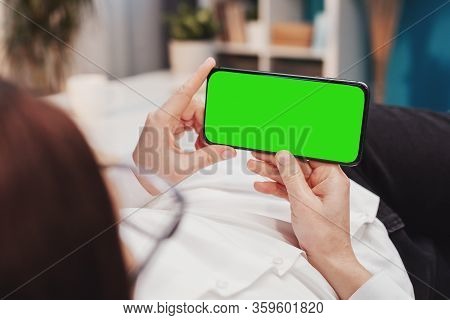 Close-up Of Human Hands Holding Horizontal Smartphone With Green Screen Lying On Bed Or Sofa Indoors