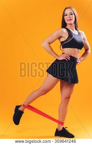 Athletic Girl Performs Exercises Using A Resistance Band. Photo Of Young Girl On Drey Background. St