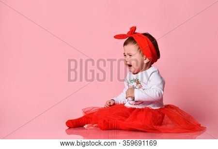 Toddler In White Bodysuit, Red Headband, Poofy Skirt, Tights. She Crying, Sitting On Floor Against P
